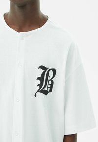Camicia bianca a maniche corte con bottoni realizzata in tessuto morbido, caratterizzata da un logo stilizzato "B" nero sulla parte sinistra del petto.