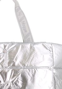 Chaqueta de color plateado metálico, acolchada, con un lazo de tela que presenta texto impreso, mostrada en un detalle de primer plano.