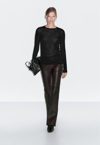 Sort tætsiddende striktrøje med lange ærmer og gennemsigtigt materiale, parret med mørkebrun læderbukser med flared fit. Modellen bærer en lille sort håndtaske.