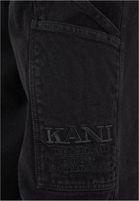 Fekete farmernadrág, nagy hímzett zsebbel, amelyen a "KANI" és a "THE ORIGINAL EST. 1989" felirat található. A textúra sima, látható varrásokkal.