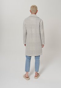 Abrigo largo en un patrón de cuadro de houndstooth en gris claro y crema, con corte recto, mangas largas y cuello con muesca, combinado con jeans azules.