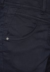 Granatowa tkanina dżinsowa z przednią kieszenią, z małym srebrnym guzikiem i kontrastowym przeplotem. Gładka faktura, minimalne detale.