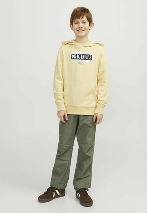 Sudadera amarilla con gráfico de "ORIGINALS", bolsillo frontal, combinada con pantalones verde oliva y zapatos marrones con rayas blancas.