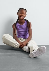 Lilla ribbet tanktopp med Nike-logo, beige joggebukser og hvite sneakers. Motivet sitter på en grå overflate mot en lys bakgrunn.