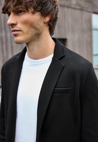 Blazer negro texturizado con solapa y un pequeño bolsillo frontal, superpuesto a una camiseta de cuello redondo blanca, en un entorno exterior desenfocado.