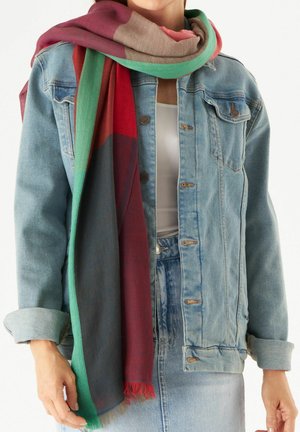 Mehrfarbiger Schal mit roten, grünen und beigen Abschnitten und Fransen, getragen über einer hellen Jeansjacke und einem weißen Oberteil.