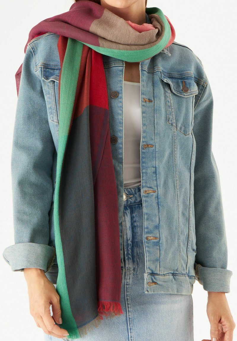 Multicolore sjaal met rode, groene en beige secties met franjes, gedragen over een lichte denimjacket en een wit topje.