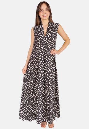 Lächelnde Frau in ärmellosem schwarzem Maxikleid mit weißen Punkten, steht mit einer Hand auf der Hüfte und Sandalen an den Füßen.