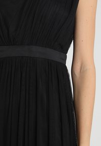 Černé šaty s plisovaným designem, jemným látkovým živůtkem a saténovým pasem. Sukně má lehkou, splývavou texturu.