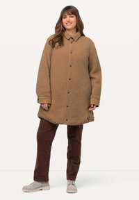 Brun fleecejacka med knappar, krage, långa ärmar och rak passform, parat med bruna kordfløyelsbyxor och ljusgrå skor.