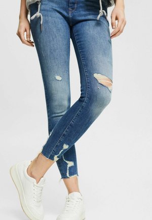 Jean skinny en denim bleu avec des zones déchirées et des ourlets effilochés, associés à des baskets blanches. Le tissu présente des nuances de bleu variées.
