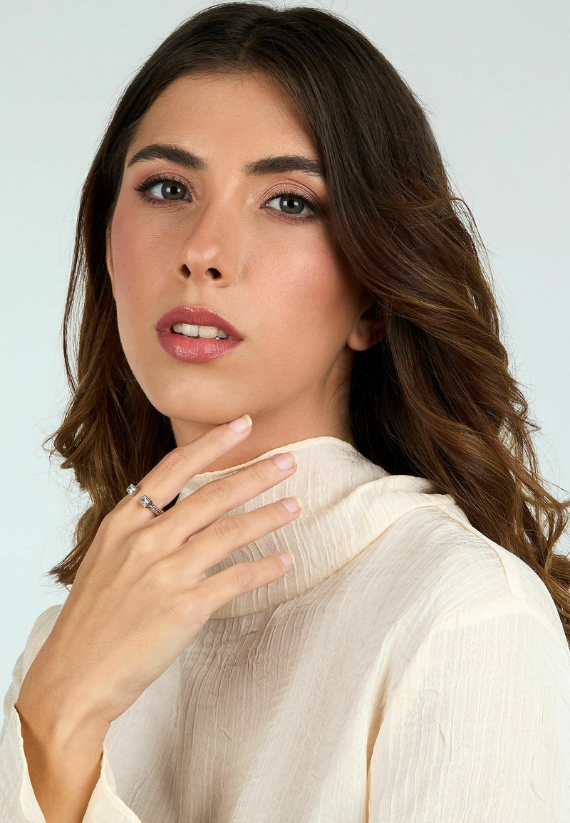 Blusa crema a maniche lunghe con colletto alto e tessuto texturizzato. Il modello indossa due anelli d'argento, mostrando unghie naturali. Sfondo semplice.