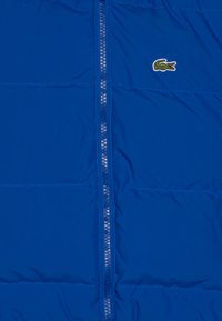 Modra napihljena jakna z navpičnim zadrgo, ki vključuje šivane dele in majhen zeleno-beli vezen kroko na levi strani.