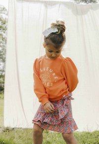 Oranje sweatshirt met puffy mouwen en bloemenmotief, gecombineerd met een meerkleurige ruches rok. Haar is vastgebonden met een witte strik.
