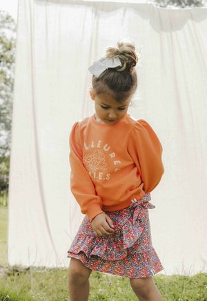 Oranje sweatshirt met puffy mouwen en bloemenmotief, gecombineerd met een meerkleurige ruches rok. Haar is vastgebonden met een witte strik.