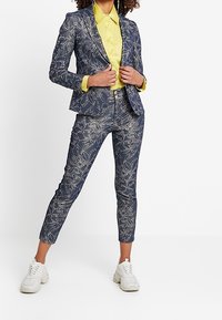 Navy-kuvioinen puku takki ja housut, joissa on valkoinen kukkakuosi, yhdistettynä keltaiseen paitaan ja valkoisiin lenkkikenkiin. Mukautettu istuvuus, tyylikäs muotoilu.