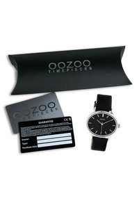Montre OOZOO avec un cadran noir, un boîtier métallique argenté et un bracelet en cuir noir. Comprend une carte de garantie noire et un emballage noir.