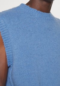 Gros plan sur une personne portant un pull sans manches, en maille duveteuse bleu clair avec des finitions côtelées au col et aux emmanchures.