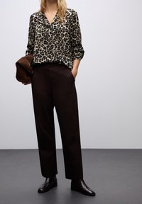 Femme portant un chemisier imprimé léopard, un pantalon marron foncé et des bottines noires, tenant une veste marron dans une main.