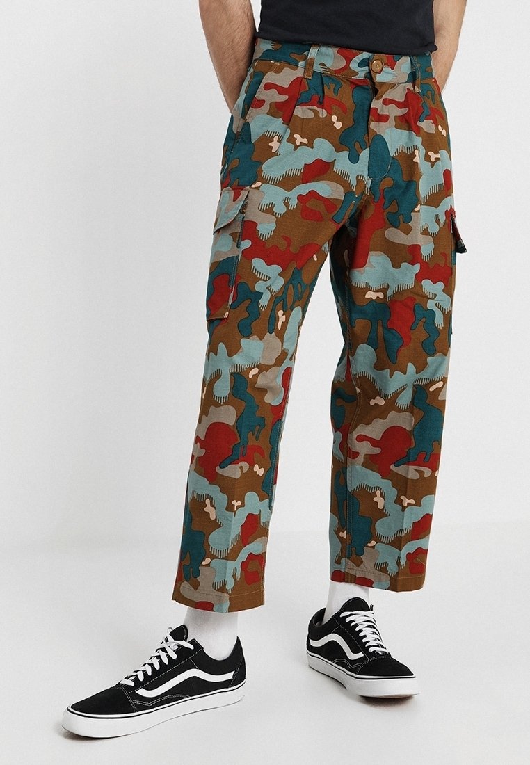 Homme portant un pantalon cargo camouflage multicolore, des chaussettes blanches et des baskets noires et blanches, debout sur un fond uni.