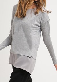 Nainen, joka on pukeutunut kerrokselliseen harmaaseen pitkähihaiseen puseroon, jossa on vaaleanharmaa sifonkialuskerros ja mustat housut, seisoo kädet hieman ojennettuina.