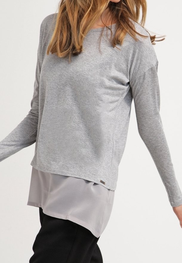 Žena v šedém dlouhém rukávu, se spodní vrstvou z lehkého šedého šifonu a černými kalhotami, stojící s mírně vystrčenýma rukama.