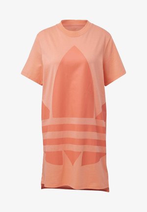 Osoba o ciemnej karnacji ubrana w luźną, brzoskwiniową sukienkę typu T-shirt z dużym logo oraz białe buty sportowe z brzoskwiniowymi akcentami.