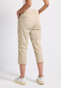 Person i beiga korta cargobyxor och vita sneakers, ståendes med händerna i bakfickorna mot en enkel vit bakgrund.