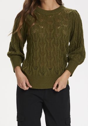 Žena v olivově zeleném pleteném svetru s texturou a nabíranými rukávy a černých kalhotách na jednobarevném pozadí.