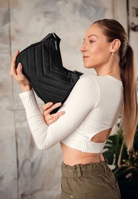 Černá prošívaná taška s chevron vzorem, se stříbrným zipem a kovovými akcenty, kterou drží modelka v zkráceném bílém topu.