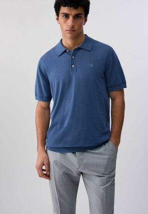 Giovane uomo che indossa una polo blu a maniche corte e pantaloni grigio chiaro, in piedi con una mano in tasca contro uno sfondo semplice.