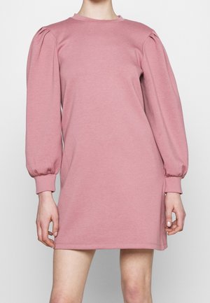 Kvinde iført en kort, støvet rosa kjole med lange puffede ærmer og tætsiddende manchetter, stående mod en ensfarvet lys baggrund.