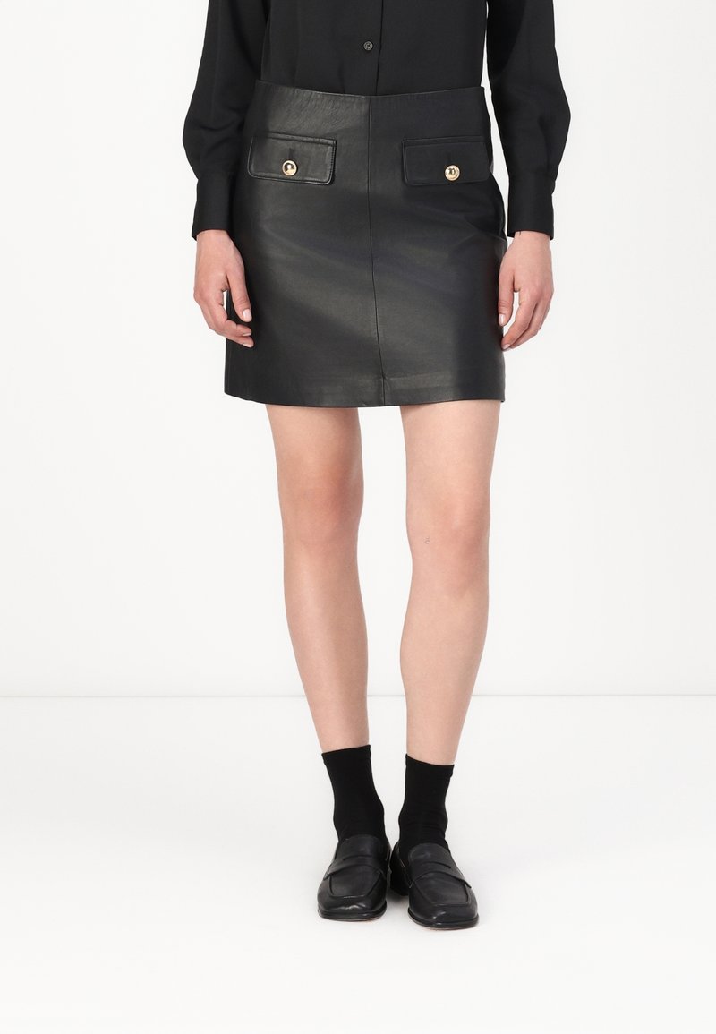 Femme portant une chemise noire à boutons, une mini-jupe en cuir noire avec poches avant, des chaussettes noires et des mocassins noirs, debout devant un fond blanc.