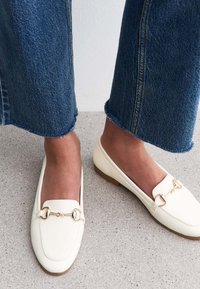 Mocassini bianchi in materiale sintetico con punta quadrata, dettagli in metallo dorato e suola piatta marrone, abbinati a jeans in denim a zampa.