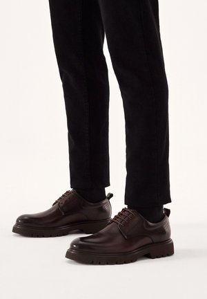 Zapatos de vestir de cuero marrón con punta redonda, diseño de cordones y suela de goma texturizada; combinados con pantalones negros ajustados.