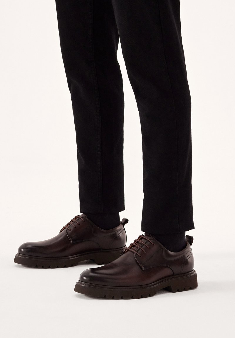 Scarpe di pelle marrone con punta arrotondata, design con lacci e suola in gomma texturizzata; abbinate a pantaloni neri a sigaretta.