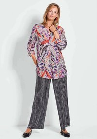 Blouse colorée à motifs avec des nuances de violet, d'orange et de noir. Associée à un pantalon large rayé noir, texturé avec de fines lignes blanches.