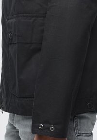 Gros plan d'une manche de veste noire et d'une poche latérale avec des détails de boutons sur un jean en denim clair.