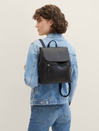 Mochila de cuero negro con cierre de solapa, superficie texturizada, bolsillo frontal con cremallera y correas ajustables, llevada sobre una chaqueta de mezclilla.