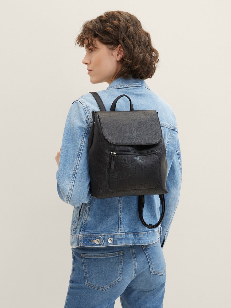 Mochila de cuero negro con cierre de solapa, superficie texturizada, bolsillo frontal con cremallera y correas ajustables, llevada sobre una chaqueta de mezclilla.