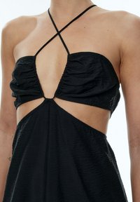 Schwarzes Kleid mit gerafften Cups, ausgeschnittenen Seiten und dünnen, kreuzförmigen Trägern. Texturiertes Material mit einer fließenden Silhouette.