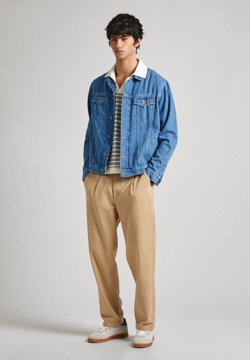 Joven de pie con las manos en los bolsillos, vistiendo una chaqueta de mezclilla azul, un suéter a rayas, pantalones beiges y zapatillas blancas sobre un fondo gris.