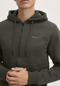 Felpa con zip di colore verde scuro con una finitura tessurizzata, dotata di cappuccio con coulisse e un piccolo logo "blend" sul petto. Tessuto liscio.