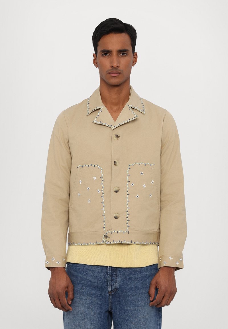 Beige cotton jacket with contrasting blue stitching, two front pockets, collar, and button closure, worn over a yellow sweater.