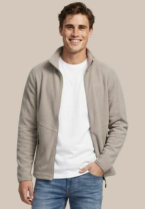 Jonge man glimlacht, draagt een lichtbeige fleecejack met rits over een wit t-shirt en blauwe spijkerbroek, staand tegen een effen achtergrond.