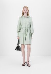 Hellgrünes Kleid mit langen Ärmeln, Knopfleiste, gerafftem Taillenbereich und strukturierten Details. Kombiniert mit einer schwarzen Handtasche und Riemchensandalen.
