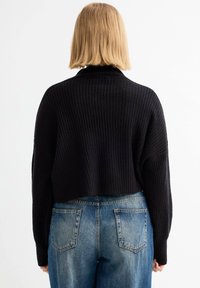 Suéter negro acanalado corto con un cuello alto, combinado con jeans de mezclilla azul claro que cuentan con bolsillos traseros y un acabado desgastado.