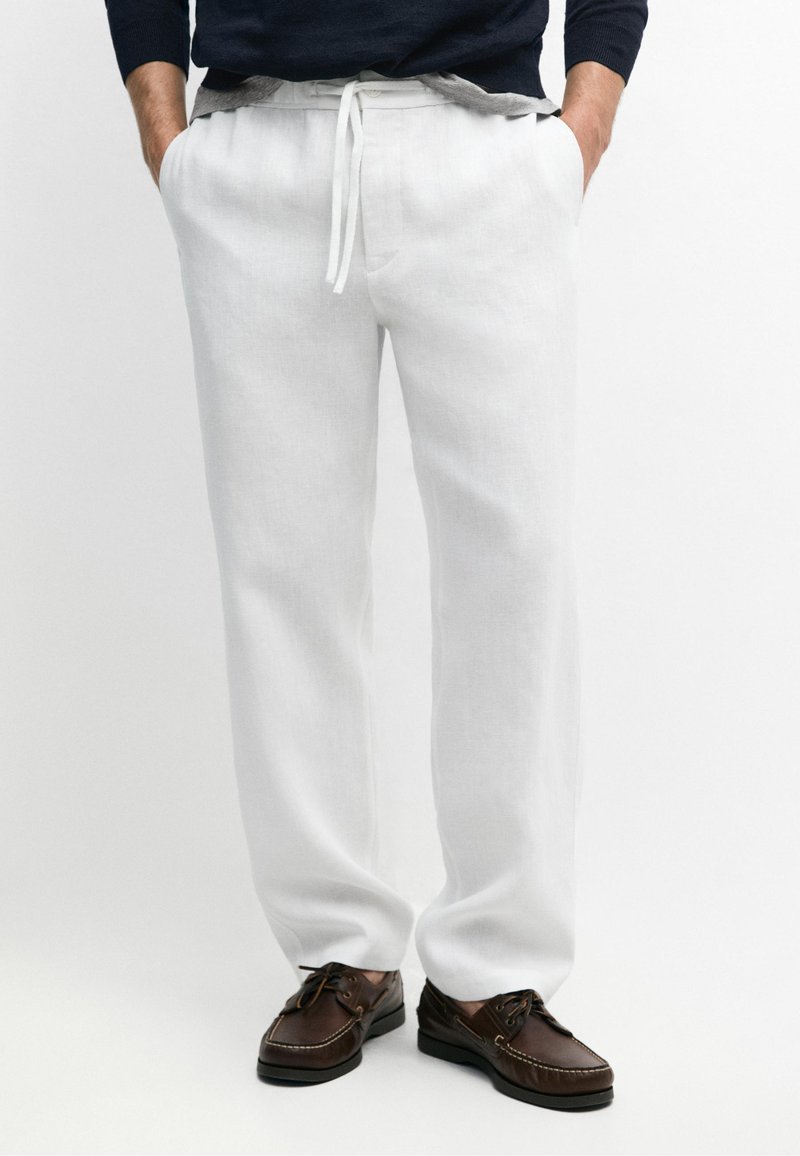 Homme portant un pantalon blanc à cordon, les mains dans les poches, chaussures bateau marron foncé sur fond uni.
