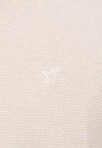 Κοντινό πλάνο σε ύφασμα κρεμ χρώματος με πλέξη βάφλας και διακριτικό κεντητό κολιμπρί στο ίδιο νήμα.