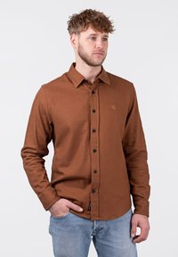 Camisa marrón de botones con cuello, que presenta botones negros y un logotipo bordado. Llevada con jeans de un azul claro.